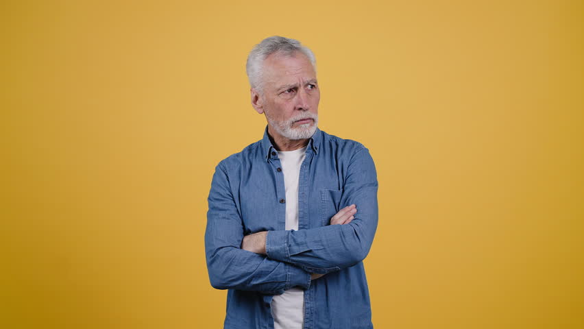 Puzzled senior man in denim shirt crosses arms and frowns looking to sides. Grey-haired pensioner thinks about solving problem on yellow background | Shutterstock HD Video #1099646165