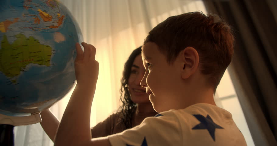 Child with mom is studying while sitting at home in rays sunset in room.Kid in morning light sunset, Cute boy Child with Globe and magnifying glass, technology for schoolboy, child looks at globe | Shutterstock HD Video #1099646917