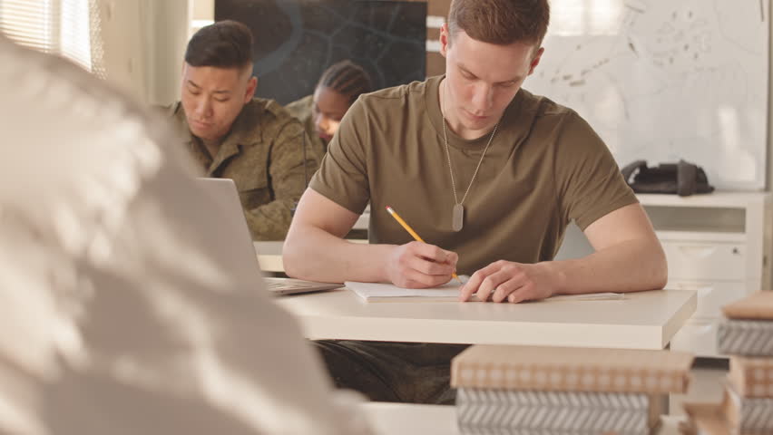Waist up slowmo of group of young multiethnic male and female cadets sitting at their desks in classroom and studying military service with professor at academy