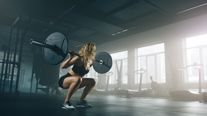 Close-up shot of a confident girl with curly, blonde hair doing squats with a barbell while exercising in the gym. Active, fit lady testing her strength with intensive workout. High quality 4k footage Royalty-Free Stock Footage #1099647523