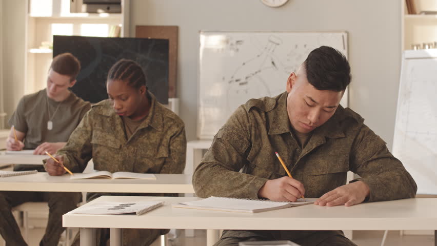 Waist up slowmo of diverse cadets sitting at desks in classroom of military academy studying