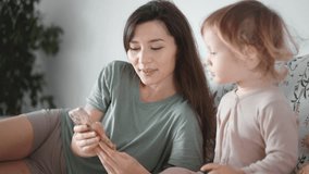 Asian young mother and curly boy daughter toddler kid look at phone screen video chat online. Woman and baby taking selfie or watch cartoons. Spend time together. Parenthood concept. bedroom morning