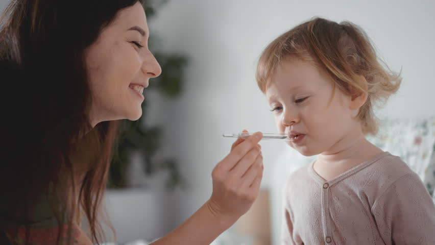 Pretty asian woman measure temperature of little girl or boy. Cute curly toddler sick at home in bedroom. Smiling kid put a thermometer at mouth. Home quarantine coronavirus. Closeup Influenza concept | Shutterstock HD Video #1099653901