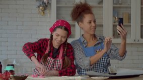 Positive pretty African American mother and lovely multiethnic teenage daughter in aprons making video call online using smartphone, talking with relatives while cooking cookies in domestic kitchen.