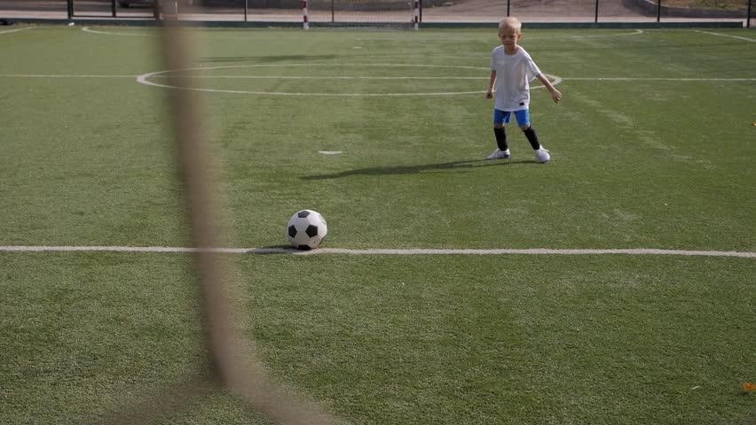 A small boy soccer player hits the goal with a soccer ball, in the foreground a blurred football net. | Shutterstock HD Video #1099655623