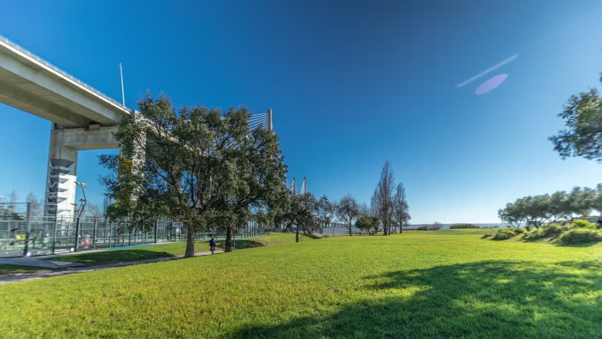 The Vasco da Gama Bridge timelapse hyperlapse viewed from green lawn with trees. Cable-stayed longest bridge flanked by viaducts and rangeviews that spans Tagus River in Park Nations. Lisbon, Portugal