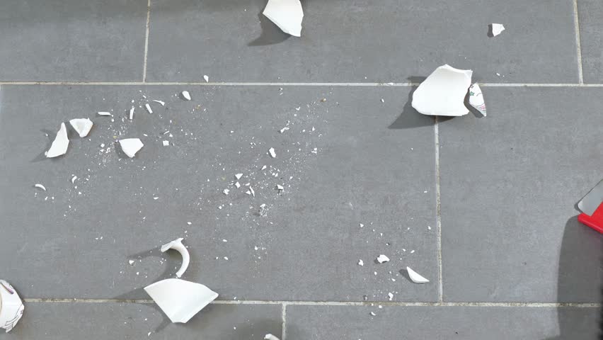 Close-up of a woman picking up fragments of a broken white ceramic mug from the floor with a red plastic scoop. The broken dishes are removed from the floor so that no one gets hurt | Shutterstock HD Video #1099669557