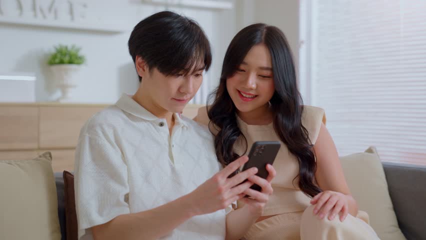 Young asian men and women watching videos on smartphones in living room on couch. Happy couple having fun using telephone at home. playing game, doing online shopping, laughing, social media apps. Royalty-Free Stock Footage #1099673531