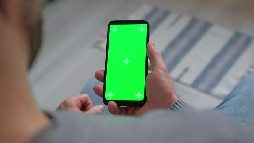 Young man sitting at home holding smartphone green mock-up screen in hand. Male person using chroma key mobile phone. Vertical mode. Touching, swiping display, tapping, surfing Internet social media | Shutterstock HD Video #1099673827
