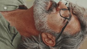 VERTICAL VIDEO: Close-up, thoughtful middle-aged man with gray hair and beard, wearing casual clothes, looking away. Mature gentleman in eyeglasses