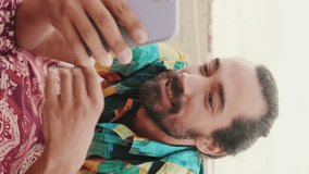 VERTICAL VIDEO: Young happy man talking by video link on mobile phone on the beach