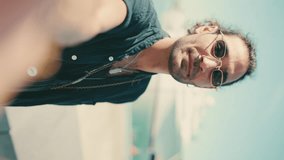 VERTICAL VIDEO: Clouse-up,young italian guy with long curly hair and stubble takes selfie on mobile phone. Stylish man in sunglasses posing smiling at camera of his phone and pointing at yacht