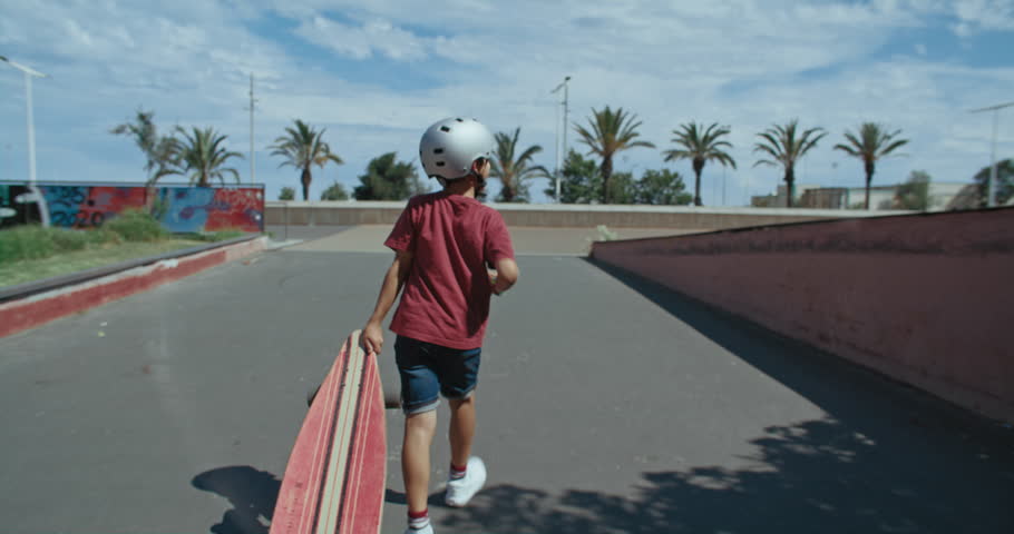 Handheld live camera follow adorable young kid in skateboarding helmet with longboard on way to skatepark on a weekend day off. Fun and active leisure activity for children Royalty-Free Stock Footage #1099679535