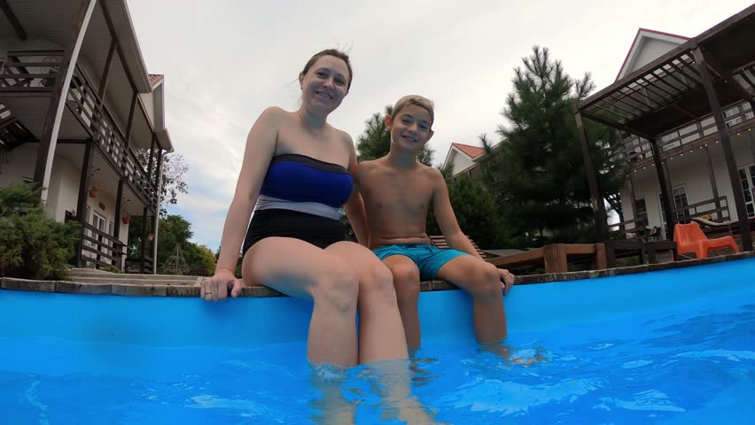Smiling boy with young mother sitting on the swimming pool talking, laughing and splashing blue water in summer day. Child and attractive woman enjoying a vacation | Shutterstock HD Video #1099681425
