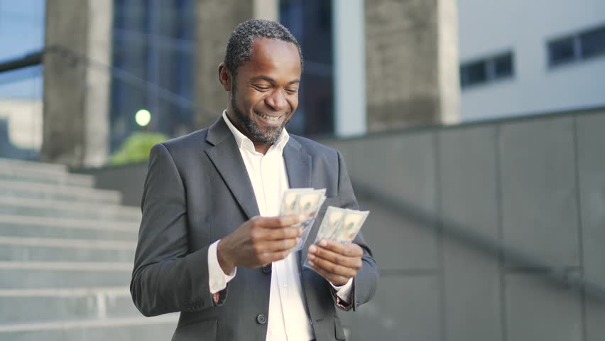 Happy african american mature man counting dollar bills outside in front of office building. A satisfied middle-aged businessman in a formal suit holds money in his hands and rejoices at good earnings | Shutterstock HD Video #1099682491