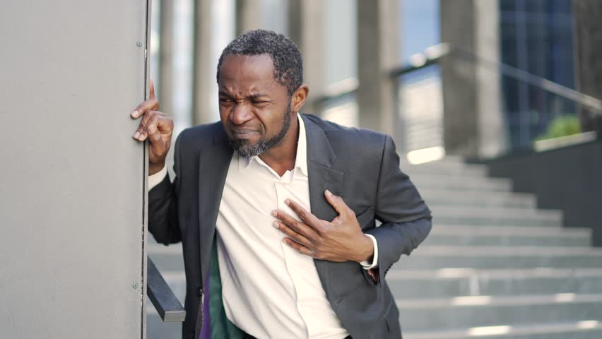 Sick african american mature man suffering from heart pain standing outside in front of office building. A middle-aged businessman in a formal suit holds his hand to his chest. He has a heart attack