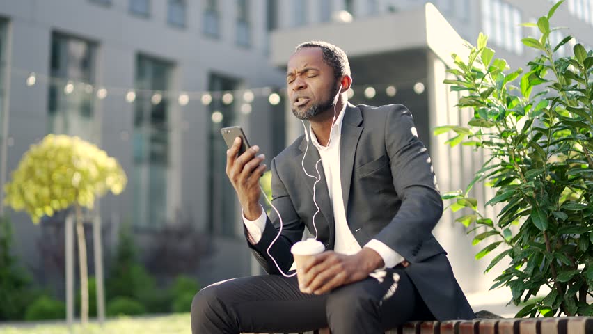 African american mature man with headphones talking on a smartphone while sitting on a bench. Businessman in a suit and holding a cup of coffee is having a conversation in front of an office building
