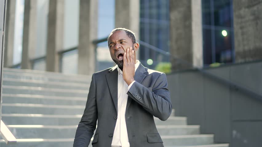 Sad african american mature man feeling toothache standing outside in front of office building. A tired middle-aged businessman in a formal suit is suffering from pain, holding his hand to his cheek | Shutterstock HD Video #1099682527