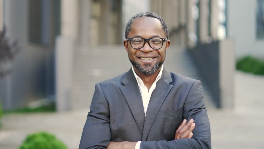 Close-up portrait of smiling mature african american man in glasses with crossed arms looking at camera outdoors. A confident businessman is standing in a formal suit in front of an office building | Shutterstock HD Video #1099682545