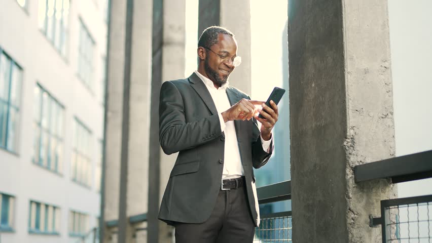 A smiling african american businessman in glasses is typing on a smartphone while standing on the street Happy mature man in a formal suit is cheerfully chatting online in front of an office building Royalty-Free Stock Footage #1099682573