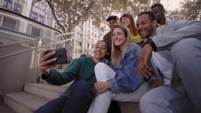 Group of young people taking a selfie or video with their phones sitting on the stairs. Smiling multiracial friends posing for a photo outside the University campus. Students in their free time.