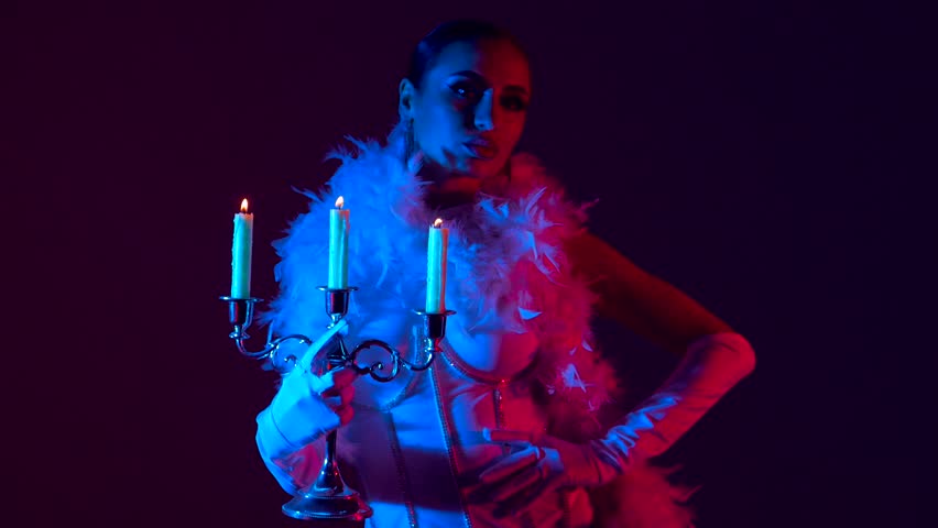 Brunette in a white dance bodysuit with a bright evening make-up with a white boa with a candelabra and three burning candles with colored red-blue lighting