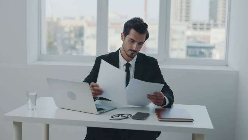Man business sits at his desk in the office with a laptop and documents and phone smile, working with online information and data processing, closing the deal. | Shutterstock HD Video #1099689481