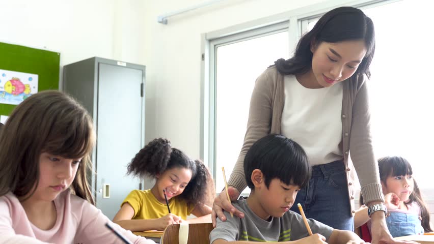 Asian school teacher assisting students in classroom. Young woman working in school helping boy with his writing, education, support, care. Royalty-Free Stock Footage #1099690991