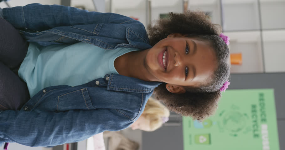 Vertical video portrait of smiling biracial schoolgirl sitting in classroom, copy space. Education, childhood, inclusivity, school and learning concept.