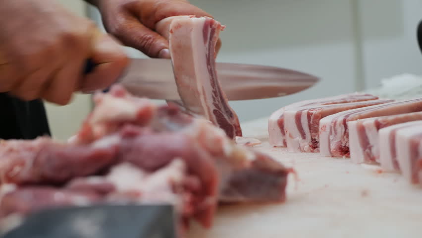 The butcher's hands are cutting bacon on a cutting table using a large sharp knife. The process of slicing pork meat with fatty layers in the meat workshop by a male worker of a meat factory | Shutterstock HD Video #1099692865