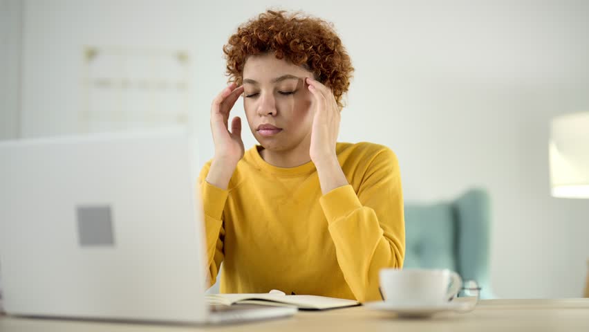 Young woman sits at computer with closed eyes and massages her temples to relieve headaches and eye fatigue after working on laptop at freelance. Tired, burnout, ill health, discomfort after work. | Shutterstock HD Video #1099697149