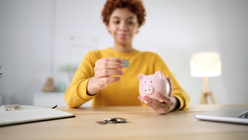 Young woman puts coins in piggy bank at home. Concept saving money, planning future investment, finance, savings, learning economy. Accounting for cash, saving money for purchases or mortgages. | Shutterstock HD Video #1099697167