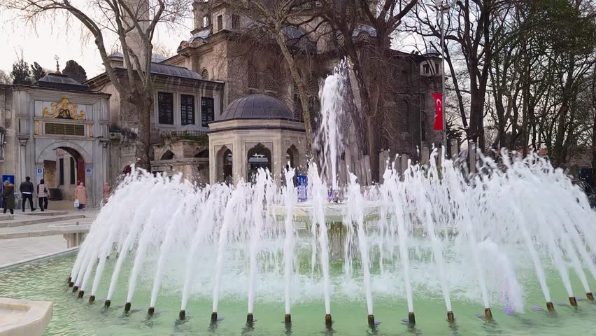  Exterior view of Eyup Sultan Camii in Istanbul. Arabic text translates  built on the grave site of Abu Ayyub al-Ansari. It is a complex building enclosed in walls. There is a pool with fountains
