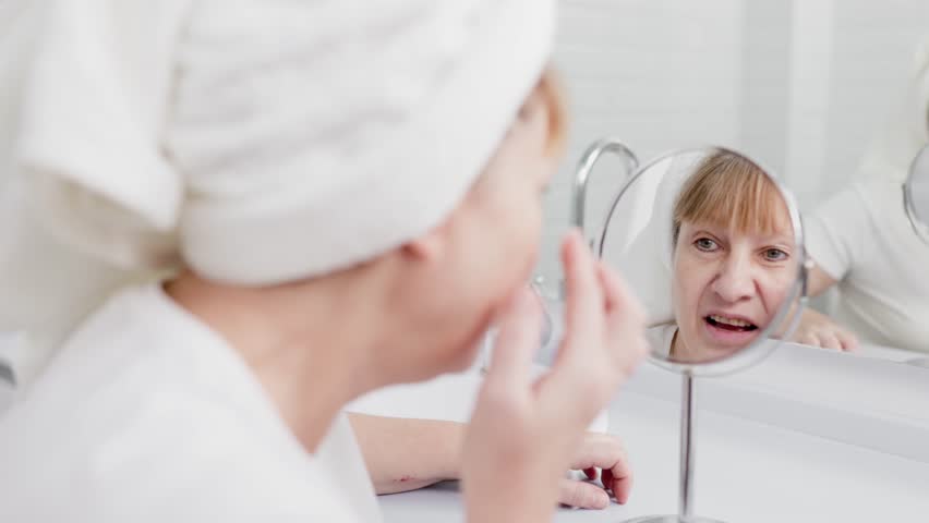 Gorgeous mid age older adult 50 years old blonde woman wears bathrobe in bathroom applying nourishing antiage face skin care cream treatment, looking at mirror doing daily morning beauty routine. Royalty-Free Stock Footage #1099700123