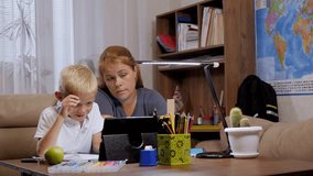An adult mother and her schoolboy son do homework during distance learning using a tablet. The child makes an entry in a notebook. Online education and e-learning concept.