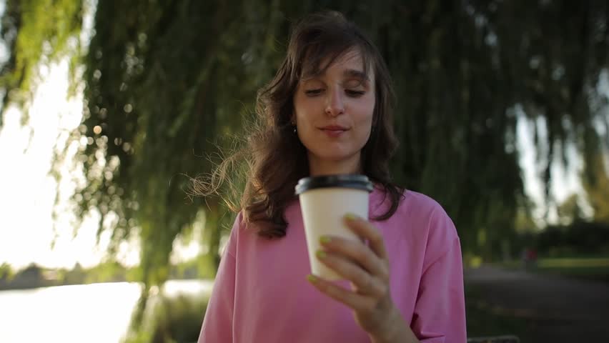 cute charming young girl in pink t-shirt, beaming smile, holding paper coffee cup, outdoor parking. Cheerful young girl in a pink t-shirt walks in the park with a cup of takeaway coffee Royalty-Free Stock Footage #1099701489