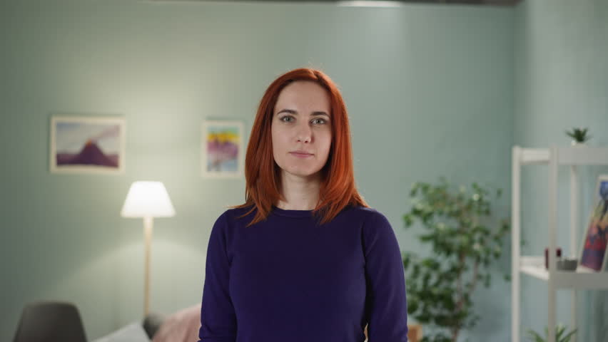 Young woman with ginger hair looks in camera tilting head to side. Portrait of positive female with smile on face standing against lamp in room closeup Royalty-Free Stock Footage #1099703479