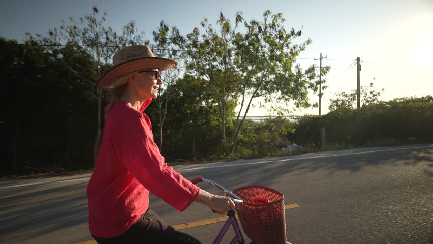 Side view portrait of pretty senior mature woman smiling wearing sunglasses, a hat, and ethnic clothes on a bicycle at sunset holding out her arm in happiness.
