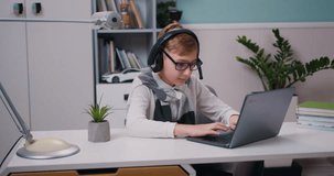 Joyful little boy in eyeglasses and headphones is playing video game using laptop sitting at his desk in living room. Gamers lifestyle.