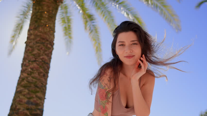 Close-up portrait of joyful young asian woman with dark hair and tattoo standing alone in seaside city smiling looking at camera. People and style concept. Palm trees ocean and mountains in background Royalty-Free Stock Footage #1099718089