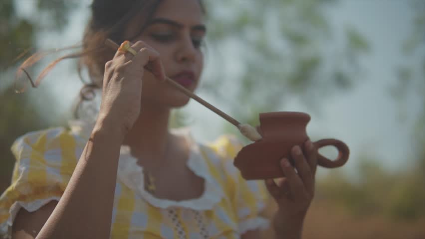 young woman standing in a field holding a stick with glue on it and applying it to a broken clay cup.