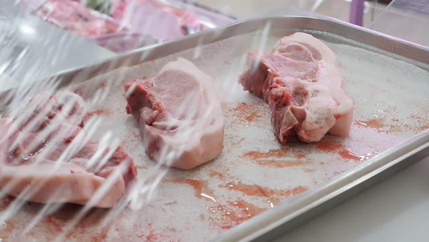 A salesman in a butcher's shop covers a tray with three raw pork entrecote with cling film. Pieces of meat in the process of trading are covered with food polyethylene according to the rules | Shutterstock HD Video #1099723343