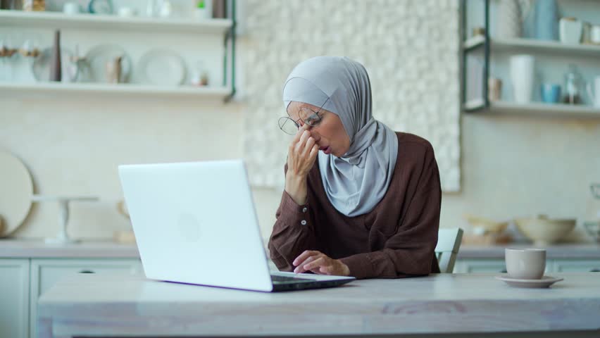 Tired muslim woman feels eye pain while working on laptop computer at home. The female freelancer is exhausted and suffering from stress Tension in glasses rubs eyes poor eyesight headache migraine | Shutterstock HD Video #1099726133