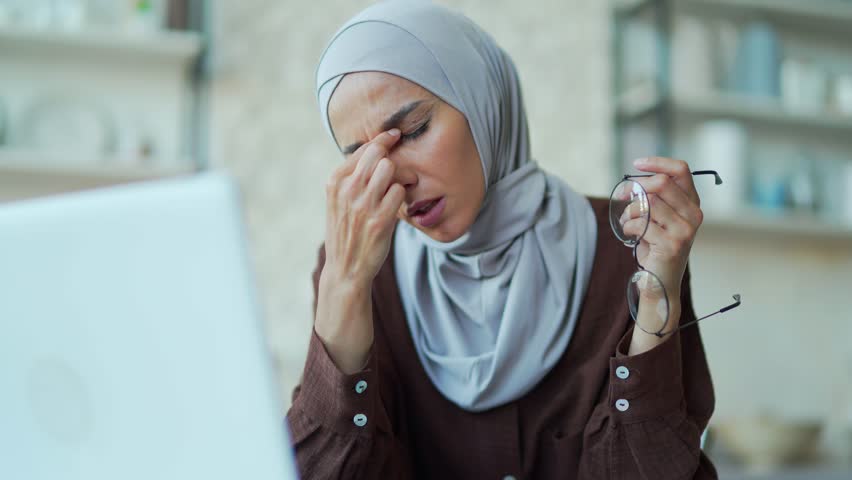 Tired muslim woman feels eye pain while working on laptop computer at home. The female freelancer is exhausted and suffering from stress Tension in glasses rubs eyes poor eyesight headache migraine | Shutterstock HD Video #1099726139