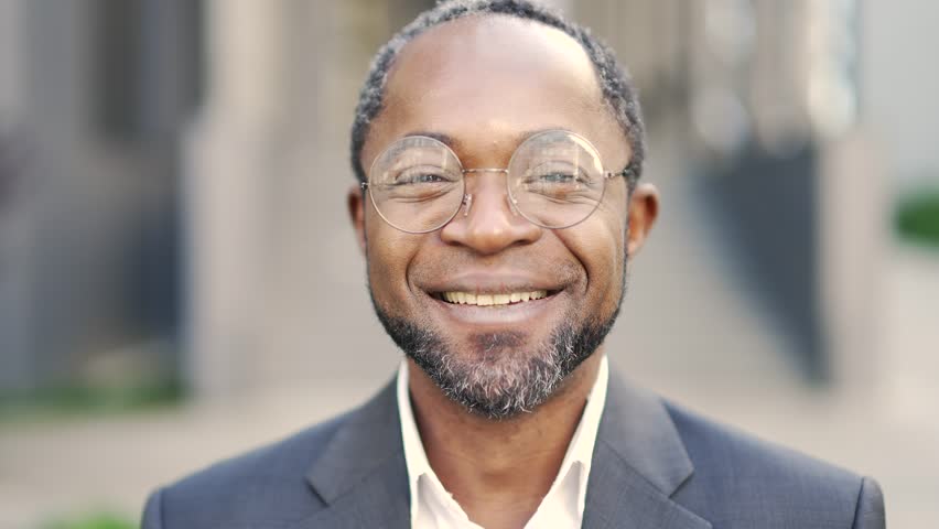 Close-up portrait of a smiling mature african american man wearing glasses looking at the camera outdoors. Сonfident middle-aged businessman is standing in a formal suit in front of an office building | Shutterstock HD Video #1099726147