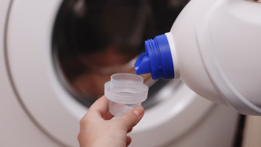 Close up shot of a woman hands squeezing liquid gel laundry detergent from a plastic bottle into a small container, with a washing machine in the background. 4k Slow motion Royalty-Free Stock Footage #1099727725