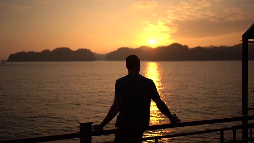 Slow motion shot, Male silhouette enjoying scenic sunset standing on pier, Sunlight reflections on water