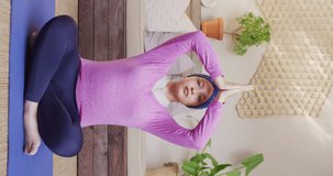 Vertical video of tranquil biracial woman in hijab sitting in yoga meditation pose at home. Happiness, health, fitness, inclusivity and domestic life.