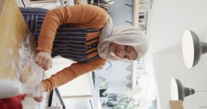 Vertical video of happy biracial woman in hijab baking in kitchen, kneading dough on worktop. Happiness, health, inclusivity and domestic life.