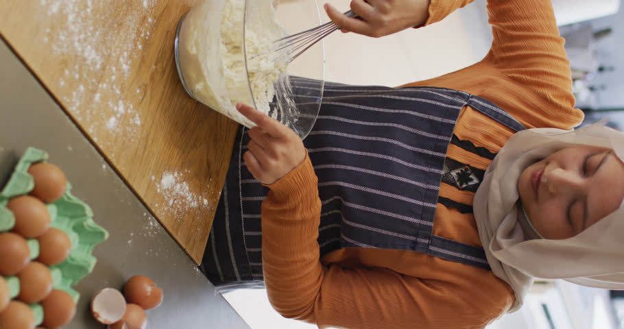 Vertical video of happy biracial woman in hijab baking in kitchen, mixing cake batter in bowl. Happiness, health, inclusivity and domestic life.